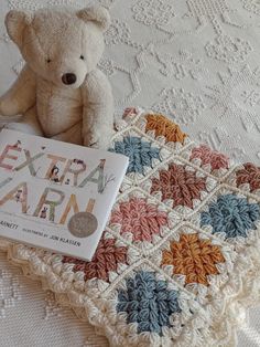 a teddy bear sitting on top of a bed next to an extra yarn book and crocheted blanket