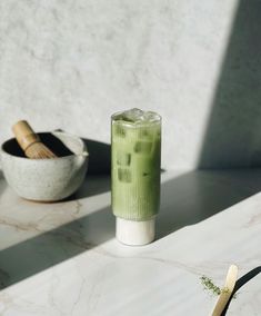 a green drink sitting on top of a white counter next to a bowl and spoon