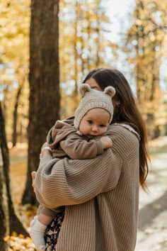 a woman holding a baby in her arms while walking through the woods with leaves on the ground