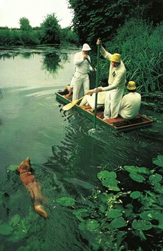 three people in a boat on the water with a dog swimming next to them,