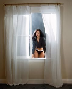 a woman standing in front of a window with sheer curtains