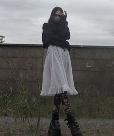 a woman in black jacket and white dress standing next to a wall with her hands on her head