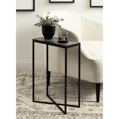 a side table with a plant on it in front of a white chair and framed pictures