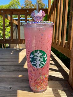 a pink starbucks drink with sprinkles and a bow on the top is sitting on a wooden deck