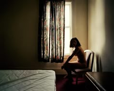 a naked woman sitting on a chair in front of a window next to a bed