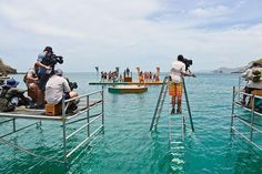 several people are sitting on small platforms in the water while others stand around watching them