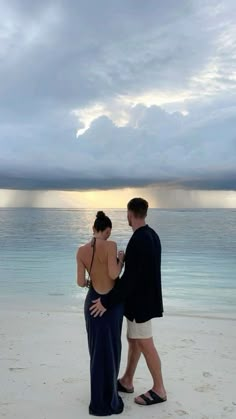 a man and woman standing on top of a beach next to the ocean under a cloudy sky