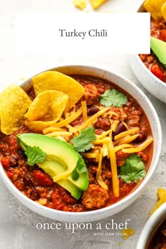 two bowls filled with chili, cheese and tortilla chips next to an avocado