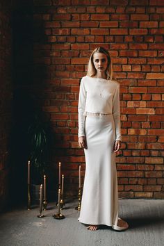 a woman standing in front of a brick wall wearing a white dress with long sleeves