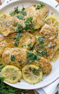 a white plate topped with chicken covered in lemon and parsley next to sliced lemons