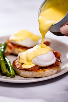 someone is pouring sauce on an english muffin with asparagus and bacon in the foreground