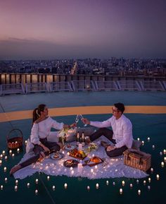 two people sitting on a blanket with food and candles in front of the city skyline