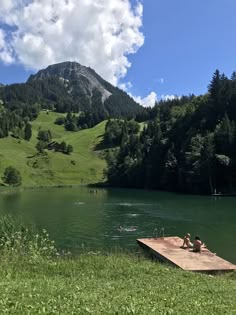 two people sitting on a dock in the middle of a lake with mountains in the background