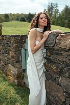 a woman in a white dress leaning against a stone wall with her hand on the shoulder