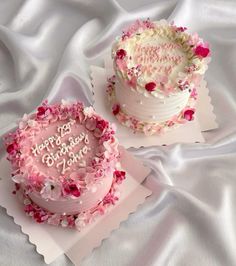 two birthday cakes with pink frosting and rose petals on white cloths, one has the words happy birthday written on it