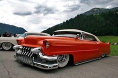 two classic cars parked next to each other in a parking lot with mountains in the background