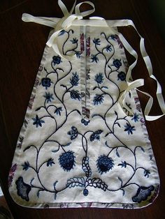 an embroidered apron with blue flowers and white ribbons on a wooden table top next to a pair of shoes