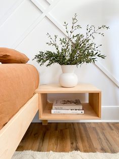 a plant in a white vase sitting on a wooden shelf next to a bed with pillows