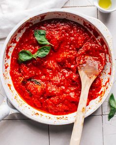 a large pot filled with sauce and basil on top of a white tile floor next to a wooden spoon