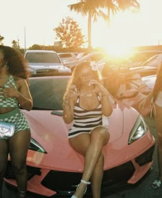 two women sitting on the hood of a pink sports car while another woman talks on her cell phone