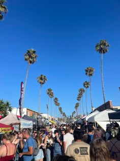 a crowd of people walking down a street next to tall palm trees and tents with signs on them