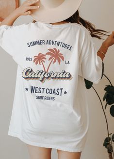 This cool retro California tshirt with palm trees design is the perfect gift idea or souvenir for every surfer in summer. Get yours now! Also available on sweatshirts: https://www.etsy.com/listing/1044743496/california-sweatshirt-los-angeles Browse through my other awesome items here: http://thenims.etsy.com/ UNISEX TEES Gildan 5000™ (CUSTOMER FAVORITE) * 100% Cotton (fiber content may vary for different colors) * Medium fabric (5.3 oz/yd² (180 g/m * Classic fit * Tear away label * Runs true to Retro Surfing T-shirt For Beach Season, Vintage Printed T-shirt For Vacation, Tropical Printed Tops For Surfing, Tropical Printed Tops For Resortwear, Summer Camp Shirt With Graphic Print For Vacation, Vacation Graphic Tee Shirt With Graphic Print, Vacation Graphic Tee With Graphic Print, Vacation Graphic Print Tee, Summer Graphic Print Camp Shirt For Vacation