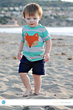 a little boy standing on top of a sandy beach next to the ocean with a fox t - shirt