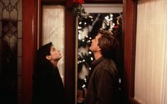 a man and woman standing in front of a mirror looking at each other with christmas lights on the window behind them