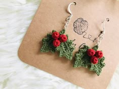 two crocheted holly leaves and red flowers hang from hooks on a piece of paper