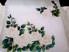 two white roses with green leaves are on a table cloth that has been embroidered onto it