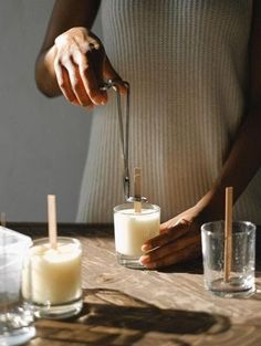 a woman is pouring some liquid into two glasses on a wooden table with candles in front of her