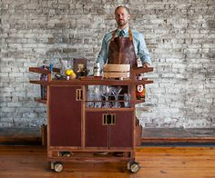 a man standing behind a bar cart with bottles on it in front of a brick wall