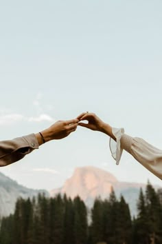 two people reaching out their hands to each other in front of mountains and pine trees