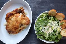 two white plates with food on them next to a bowl of salad and some bread