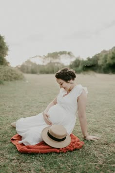 a pregnant woman in a white dress and hat sitting on a red blanket with her legs crossed