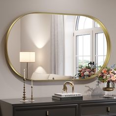 a mirror on the wall above a dresser with flowers and a lamp next to it