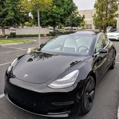 a black tesla parked in a parking lot next to some trees and cars on the street