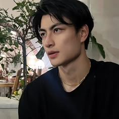 a young man with black hair wearing a black shirt and gold necklace, sitting in front of a potted plant