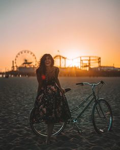 a woman standing next to a bike on top of a sandy beach at sunset with ferris wheel in the background