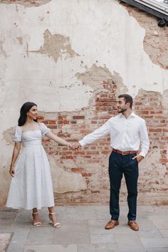 a man and woman standing next to each other holding hands in front of a brick wall