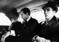 black and white photograph of two men sitting in the back seat of a car, one wearing a hat