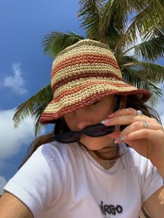 a woman wearing a hat and sunglasses is holding her hand up to her face while standing under a palm tree