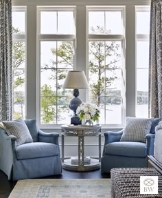 a living room with two chairs and a table in front of the large windows that have curtains on them