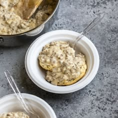 two white bowls filled with biscuits covered in gravy next to a silver pot