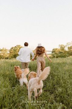 a man and woman walking with two dogs in the grass, while another dog runs behind them