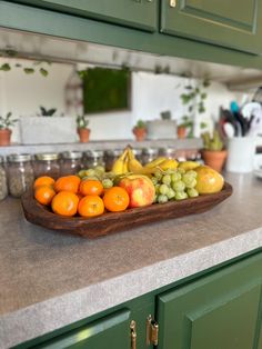 a wooden tray filled with lots of fruit on top of a counter next to green cabinets