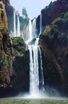 a large waterfall in the middle of a body of water surrounded by rocks and greenery