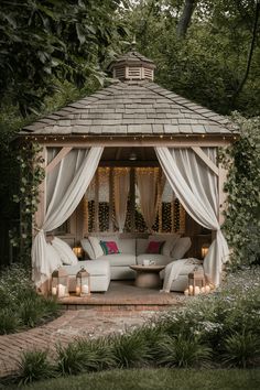 a gazebo with curtains and lights in the middle of a garden area surrounded by greenery