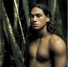 a man with long hair and no shirt standing in front of some trees looking at the camera