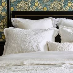 a white bed topped with pillows next to a wallpapered headboard and night stand